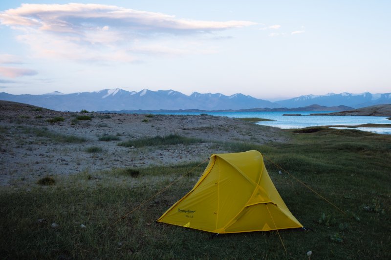 Место стоянки мне понравилось – вода,
удобное место под палатку, боковой овраг, в котором можно спокойно спрятаться
от западного ветра и приготовить еду. Спокойно поужинав, я с наступлением
темноты улегся спать.
День
5 (18.08.2025) 19 км.
Проснулся в 5:30. Вершины Заалайского
хребта, обычно хорошо видимые утром, сегодня были закрыты облаками. За ночь
уровень воды в реке упал более чем вдвое, течение стало спокойным, а вода
прозрачной. Позавтракав, заполнил все пустые бутылки водой. Теперь у меня было девять
литров воды, пригодной для питья, пусть и после кипячения. Такой запас позволял
мне гарантированно дойти до поселка.
На воду вышел в 9:08, проведя
байдарку по устью через мели. Вода в реке утром жутко холодная, прямо обжигала
ноги.
Еще не успел дойти до оконечности
первого по пути мыса, как задул сильный встречный ветер, поднялась приличная
волна. Но самое неприятное заключалось в том, что солнце пропало. Тучи с Заалайского
хребта позли навстречу и закрыли небо над озером. Стало холодно, брызги со
встречных волн обдавали меня, а вышел я как обычно в полипропиленовой футболке
с длинным рукавом. Тешил себя, что согреюсь от усиленной гребли.
Выйдя за мыс, увидел такую картину - с
долины Маркансу в долину, образованную дельтой реки Караджилга, над землей ползло
облако. Небо с северо-западной стороны тоже было затянуто кучевыми облаками,
выглядело все мрачно. Но надеясь, что позже распогодится, продолжил движение
дальше.
Пройдя первый залив, у мыса подвернул
вправо, чтобы по кратчайшему пути дойти к северному берегу следующего залива. Ветер
все продолжал усиливаться и был сейчас четко в лицо. Во время его порыва скорость
байдарки падала почти до нулевой, если не до отрицательной, но главное - удавалось
сохранять направление, не допуская поворота байдарки боком к волне. Мысли были
в этот момент только о том, чтобы ветром не вырвало из рук весло, пальцы уже сильно
замерзли.
Дошел в 10:03 до обрывистого
северного берега, здесь в зоне тени смог вылезти на прибрежные камни, поддуть
борта байдарки и съесть пару конфеток-халвы. Без ветра немного согрелся, ситуация
выглядела вполне терпимой, несмотря и на отсутствие солнца. Это привело к серьезной
ошибке – я не надел куртку и двинулся вдоль берега на восток.
У края мыса, отделяющего от
следующего залива, увидел, что теперь уже вся долина дельты Караджилги затянута
облаком, берег был плохо различим. Судя по GPS, до следующего мыса было метров
300-400, поэтому погреб дальше, несмотря на сильный боковой ветер и периодически
обливавшие слева волны. Дойдя до этого мыса, обнаружил, что пристать к берегу
не получится, крайне полого, мели начинались за несколько десятков метров от
берега. Проверил веслом дно - топкое. В тумане дальше на берегу проглядывались какие-то
стоящие многометровые фигуры, похожие то на средневековые строения, то на
сказочных чудищ. Это были образованные выветриванием останцы, как и на юге, но сейчас
они выглядели устрашающе.
Теперь требовалось пересечь вытянутый
на север на несколько километров залив, берег которого образован дельтой
Караджилги. В поселке меня предостерегали от высадки в тех местах, предупреждая
о зыбунах. У кочевников неоднократно бесследно исчезал скот в этом районе, сейчас
они воздерживаются от выпаса там. Поэтому в залив заходить я не стал, а направился
напрямую к его восточному берегу. Пока догреб до него – начало поколачивать от
холода. Сильный ветер, брызги с весла и волн, а главное - отсутствие солнца
делали свое дело. Повернув направо, двинулся к оконечности мыса, серфя на
волне. Периодически пересекал тянущиеся от берега на запад мели, становилось
совсем неприятно – волна на них ломается, гребни накрывают байдарку то с одного
бока, то с другого. Юбка хоть и нейлоновая, но тоже немного пропускает воду. Я
все больше замерзал. Пальцев рук уже не чувствовал, хотя весло они еще держали.
Завернув за следующий мыс, решил -
надо приставать во что бы то ни стало. В 11:30 сумел найти мель с относительно
твердым дном и вылез метрах в двадцати от берега, до которого уже провел байдарку.
Еле гнущимися от холода руками снял всю мокрую одежду, натянул полартек, сверху
мембранный костюм, а поверх него для тепла спасжилет. Взяв воду, сухари и
конфеты, пошел по берегу искать хоть какое-то укрытие от ветра. Отойдя метров
двести, нашел подходящее место, за одним из редких на этом мысе камней. Сидя
спиной к нему, можно было немного спрятаться от ветра и обдумать планы на
будущее.
Довольно скоро я согрелся и решил,
что все не так уж и плохо. Непогода не может продолжаться бесконечно, сидеть
здесь безопасно, запасов воды и еды у меня дня на два минимум. Пока буду сидеть
и наблюдать за погодой.
Через пару часов из-за
туч вышло солнце, стало гораздо веселее. С Заалайского хребта тучи больше не
тянуло, ветер поменялся на WNW и, кажется, начал ослабевать. Кучевые облака теперь
клубились над хребтом Каракуль, оттуда они постепенно уходили на восток. Облако
тумана, стоявшее над дельтой реки Караджилга исчезло. Еще через час небо стало
почти чистым от облаков, настроение улучшилось, и я немного прогулялся по мысу,
пофотографировал. Место стоянки мне понравилось – вода,
удобное место под палатку, боковой овраг, в котором можно спокойно спрятаться
от западного ветра и приготовить еду. Спокойно поужинав, я с наступлением
темноты улегся спать.
День
5 (18.08.2025) 19 км.
Проснулся в 5:30. Вершины Заалайского
хребта, обычно хорошо видимые утром, сегодня были закрыты облаками. За ночь
уровень воды в реке упал более чем вдвое, течение стало спокойным, а вода
прозрачной. Позавтракав, заполнил все пустые бутылки водой. Теперь у меня было девять
литров воды, пригодной для питья, пусть и после кипячения. Такой запас позволял
мне гарантированно дойти до поселка.
На воду вышел в 9:08, проведя
байдарку по устью через мели. Вода в реке утром жутко холодная, прямо обжигала
ноги.
Еще не успел дойти до оконечности
первого по пути мыса, как задул сильный встречный ветер, поднялась приличная
волна. Но самое неприятное заключалось в том, что солнце пропало. Тучи с Заалайского
хребта позли навстречу и закрыли небо над озером. Стало холодно, брызги со
встречных волн обдавали меня, а вышел я как обычно в полипропиленовой футболке
с длинным рукавом. Тешил себя, что согреюсь от усиленной гребли.
Выйдя за мыс, увидел такую картину - с
долины Маркансу в долину, образованную дельтой реки Караджилга, над землей ползло
облако. Небо с северо-западной стороны тоже было затянуто кучевыми облаками,
выглядело все мрачно. Но надеясь, что позже распогодится, продолжил движение
дальше.
Пройдя первый залив, у мыса подвернул
вправо, чтобы по кратчайшему пути дойти к северному берегу следующего залива. Ветер
все продолжал усиливаться и был сейчас четко в лицо. Во время его порыва скорость
байдарки падала почти до нулевой, если не до отрицательной, но главное - удавалось
сохранять направление, не допуская поворота байдарки боком к волне. Мысли были
в этот момент только о том, чтобы ветром не вырвало из рук весло, пальцы уже сильно
замерзли.
Дошел в 10:03 до обрывистого
северного берега, здесь в зоне тени смог вылезти на прибрежные камни, поддуть
борта байдарки и съесть пару конфеток-халвы. Без ветра немного согрелся, ситуация
выглядела вполне терпимой, несмотря и на отсутствие солнца. Это привело к серьезной
ошибке – я не надел куртку и двинулся вдоль берега на восток.
У края мыса, отделяющего от
следующего залива, увидел, что теперь уже вся долина дельты Караджилги затянута
облаком, берег был плохо различим. Судя по GPS, до следующего мыса было метров
300-400, поэтому погреб дальше, несмотря на сильный боковой ветер и периодически
обливавшие слева волны. Дойдя до этого мыса, обнаружил, что пристать к берегу
не получится, крайне полого, мели начинались за несколько десятков метров от
берега. Проверил веслом дно - топкое. В тумане дальше на берегу проглядывались какие-то
стоящие многометровые фигуры, похожие то на средневековые строения, то на
сказочных чудищ. Это были образованные выветриванием останцы, как и на юге, но сейчас
они выглядели устрашающе.
Теперь требовалось пересечь вытянутый
на север на несколько километров залив, берег которого образован дельтой
Караджилги. В поселке меня предостерегали от высадки в тех местах, предупреждая
о зыбунах. У кочевников неоднократно бесследно исчезал скот в этом районе, сейчас
они воздерживаются от выпаса там. Поэтому в залив заходить я не стал, а направился
напрямую к его восточному берегу. Пока догреб до него – начало поколачивать от
холода. Сильный ветер, брызги с весла и волн, а главное - отсутствие солнца
делали свое дело. Повернув направо, двинулся к оконечности мыса, серфя на
волне. Периодически пересекал тянущиеся от берега на запад мели, становилось
совсем неприятно – волна на них ломается, гребни накрывают байдарку то с одного
бока, то с другого. Юбка хоть и нейлоновая, но тоже немного пропускает воду. Я
все больше замерзал. Пальцев рук уже не чувствовал, хотя весло они еще держали.
Завернув за следующий мыс, решил -
надо приставать во что бы то ни стало. В 11:30 сумел найти мель с относительно
твердым дном и вылез метрах в двадцати от берега, до которого уже провел байдарку.
Еле гнущимися от холода руками снял всю мокрую одежду, натянул полартек, сверху
мембранный костюм, а поверх него для тепла спасжилет. Взяв воду, сухари и
конфеты, пошел по берегу искать хоть какое-то укрытие от ветра. Отойдя метров
двести, нашел подходящее место, за одним из редких на этом мысе камней. Сидя
спиной к нему, можно было немного спрятаться от ветра и обдумать планы на
будущее.
Довольно скоро я согрелся и решил,
что все не так уж и плохо. Непогода не может продолжаться бесконечно, сидеть
здесь безопасно, запасов воды и еды у меня дня на два минимум. Пока буду сидеть
и наблюдать за погодой.
Через пару часов из-за
туч вышло солнце, стало гораздо веселее. С Заалайского хребта тучи больше не
тянуло, ветер поменялся на WNW и, кажется, начал ослабевать. Кучевые облака теперь
клубились над хребтом Каракуль, оттуда они постепенно уходили на восток. Облако
тумана, стоявшее над дельтой реки Караджилга исчезло. Еще через час небо стало
почти чистым от облаков, настроение улучшилось, и я немного прогулялся по мысу,
пофотографировал.