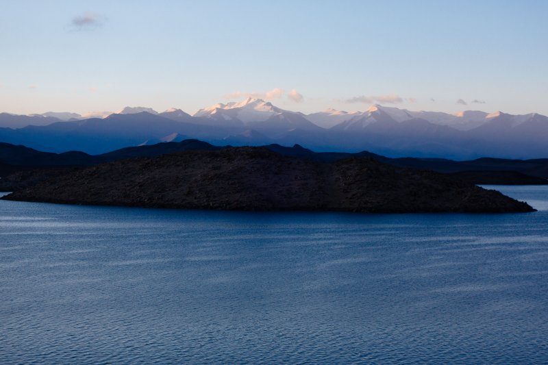 На спуске с холма нашел удутую вчера от
меня в темноте ветром флисовую перчатку, что еще больше улучшило настроение. Я-то
с ней уже распрощался.
Позавтракав и собрав вещи, принялся
за ремонт байдарки. Поверхность пвх-ткани вокруг пробоины очистил от пыли и
влаги ватным диском, смоченным ацетоном. Потом залепил пробоину двумя слоями крест-накрест
сантехнического скотча с каждой стороны. На дырку в баллоне я пожалел скотч,
налепив только один слой. И зря! Надув борта убедился, что скотч тут же начал
пропускать воздух. Ну да и ладно, буду
поддувать как раньше, оставалось немного - по карте до поселка чуть более
двадцати километров. Хотелось удрать побыстрее, пока штиль.
Однако все равно провозился долго,
из-за помороженных вчера пальцев. Дистальные фаланги опухли и приобрели
красноватый с синим оттенком цвет. Было больно прикасаться ко всему. Как
результат - стартовал только в 9:47, нацелившись сначала на восточный край
залива за полуостровом, где находился. Приблизившись к берегу, пошел вдоль него
на юго-восток. Помня о вчерашнем происшествии с мелью, аккуратно прошел между
двумя островами, перепугав сидевших там местных чаек. Чем они тут питаются – загадка, рыбы то в
озере нет. Хотя кто-то писал, что якобы в заливах у устьев самых крупных рек,
впадающих в озеро – Музкола, Акджилги и Караджилги, вода более пресная и там водится
мелкий голец.
В какой-то момент я повернулся направо
и поразился увиденному, в небе над берегом, напротив северной оконечности
хребта Каракуль, стояло кучевое облако, а до него от земли тянулся расширяющийся
вверх белый столб. Столб медленно двигался и изгибался. Торнадо, кошмар! В
интернете была информация про частые торнадо в долинах севернее озера. Похоже
не даром одно из возможных значений топонима «Маркансу» - долина смерчей. Даже
не предполагал, что увижу такое явление. Канадец, бывший здесь на каяке за год
до меня, также видел торнадо приблизительно там же, и успел заснять на видео. Самое
интересное, что в той части озера, где я находился в этот момент, было все
прекрасно, дул легкий попутный ветерок. Что есть мочи погреб дальше к поселку,
который виднелся вдали. Через некоторое время запыхался и повернулся посмотреть,
где сейчас торнадо, но его уже не было. На всякий случай продолжал усиленно грести,
стараясь отойти на максимальное расстояние от севера озера. Вылез на берег поддуть
борта только в 11:55, до поселка оставалось совсем немного - семь километров. На спуске с холма нашел удутую вчера от
меня в темноте ветром флисовую перчатку, что еще больше улучшило настроение. Я-то
с ней уже распрощался.
Позавтракав и собрав вещи, принялся
за ремонт байдарки. Поверхность пвх-ткани вокруг пробоины очистил от пыли и
влаги ватным диском, смоченным ацетоном. Потом залепил пробоину двумя слоями крест-накрест
сантехнического скотча с каждой стороны. На дырку в баллоне я пожалел скотч,
налепив только один слой. И зря! Надув борта убедился, что скотч тут же начал
пропускать воздух. Ну да и ладно, буду
поддувать как раньше, оставалось немного - по карте до поселка чуть более
двадцати километров. Хотелось удрать побыстрее, пока штиль.
Однако все равно провозился долго,
из-за помороженных вчера пальцев. Дистальные фаланги опухли и приобрели
красноватый с синим оттенком цвет. Было больно прикасаться ко всему. Как
результат - стартовал только в 9:47, нацелившись сначала на восточный край
залива за полуостровом, где находился. Приблизившись к берегу, пошел вдоль него
на юго-восток. Помня о вчерашнем происшествии с мелью, аккуратно прошел между
двумя островами, перепугав сидевших там местных чаек. Чем они тут питаются – загадка, рыбы то в
озере нет. Хотя кто-то писал, что якобы в заливах у устьев самых крупных рек,
впадающих в озеро – Музкола, Акджилги и Караджилги, вода более пресная и там водится
мелкий голец.
В какой-то момент я повернулся направо
и поразился увиденному, в небе над берегом, напротив северной оконечности
хребта Каракуль, стояло кучевое облако, а до него от земли тянулся расширяющийся
вверх белый столб. Столб медленно двигался и изгибался. Торнадо, кошмар! В
интернете была информация про частые торнадо в долинах севернее озера. Похоже
не даром одно из возможных значений топонима «Маркансу» - долина смерчей. Даже
не предполагал, что увижу такое явление. Канадец, бывший здесь на каяке за год
до меня, также видел торнадо приблизительно там же, и успел заснять на видео. Самое
интересное, что в той части озера, где я находился в этот момент, было все
прекрасно, дул легкий попутный ветерок. Что есть мочи погреб дальше к поселку,
который виднелся вдали. Через некоторое время запыхался и повернулся посмотреть,
где сейчас торнадо, но его уже не было. На всякий случай продолжал усиленно грести,
стараясь отойти на максимальное расстояние от севера озера. Вылез на берег поддуть
борта только в 11:55, до поселка оставалось совсем немного - семь километров.
